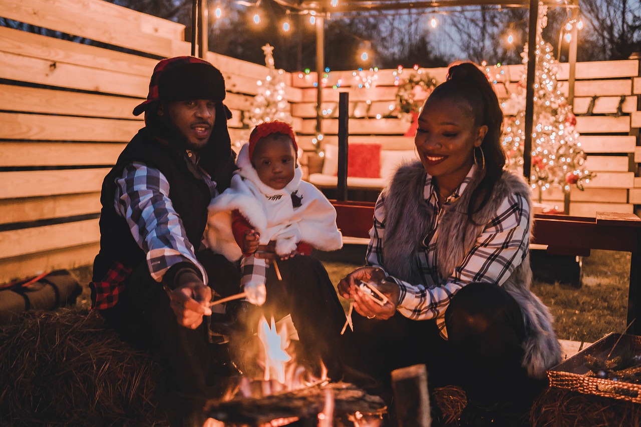 A mother and father sitting by a campfire in the evening, with their 3-year-old child sitting on the father’s lap, looking at the fire while he roasts a marshmallow. The family is smiling and enjoying a cozy moment together.