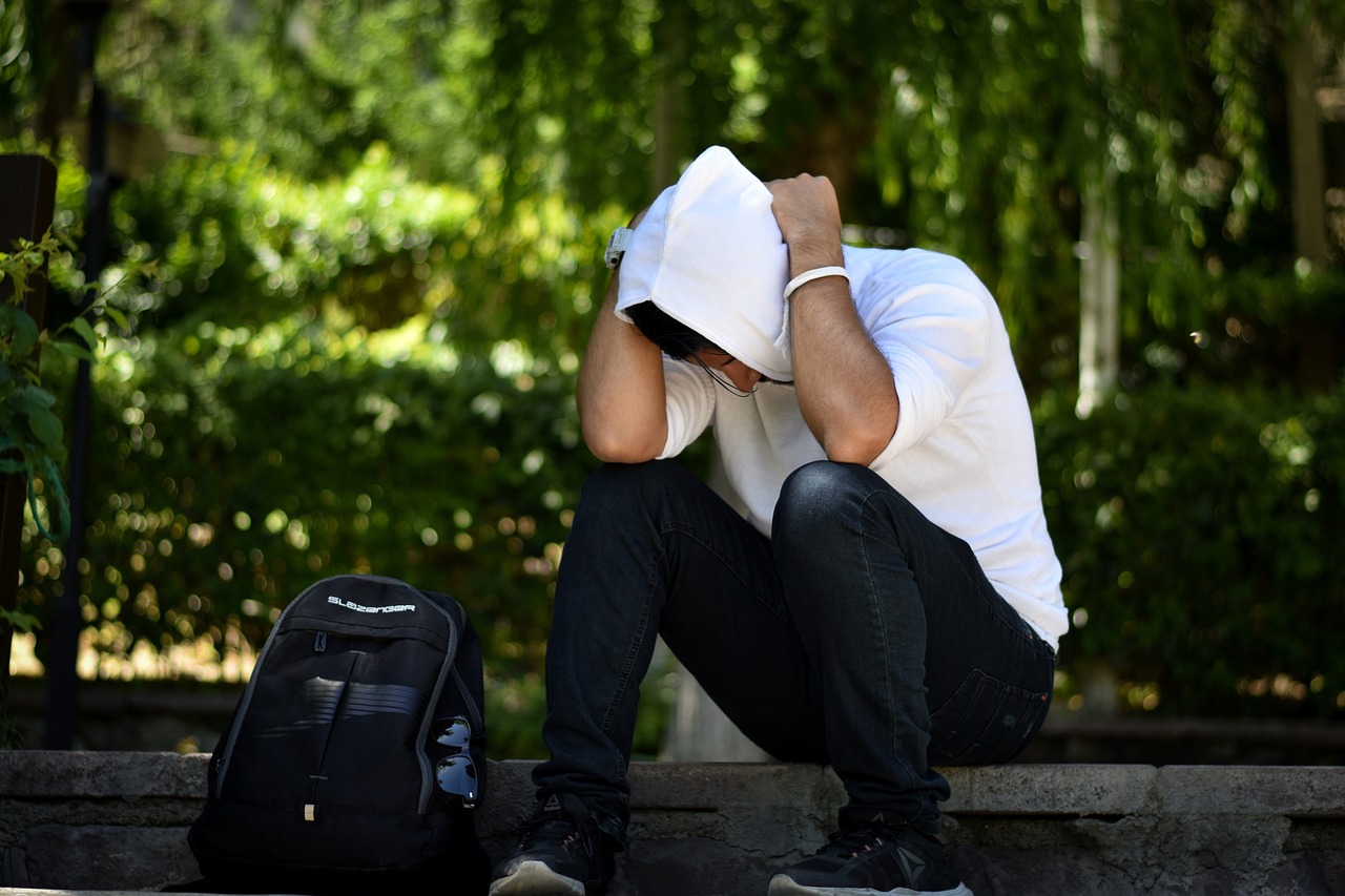 Man sitting on a stoop along a walking trail, wearing a white hoodie with the hood up, head bowed and hands clasped behind his head, looking down with a stressed expression, reflecting a moment of overwhelm.