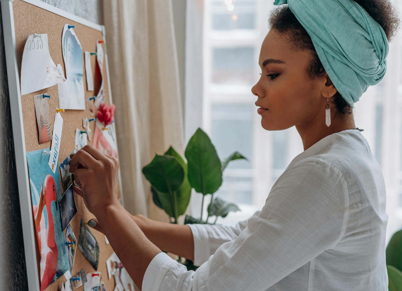 A woman standing in front of a vision board, arranging photos and quotes on a corkboard in a bright home office.